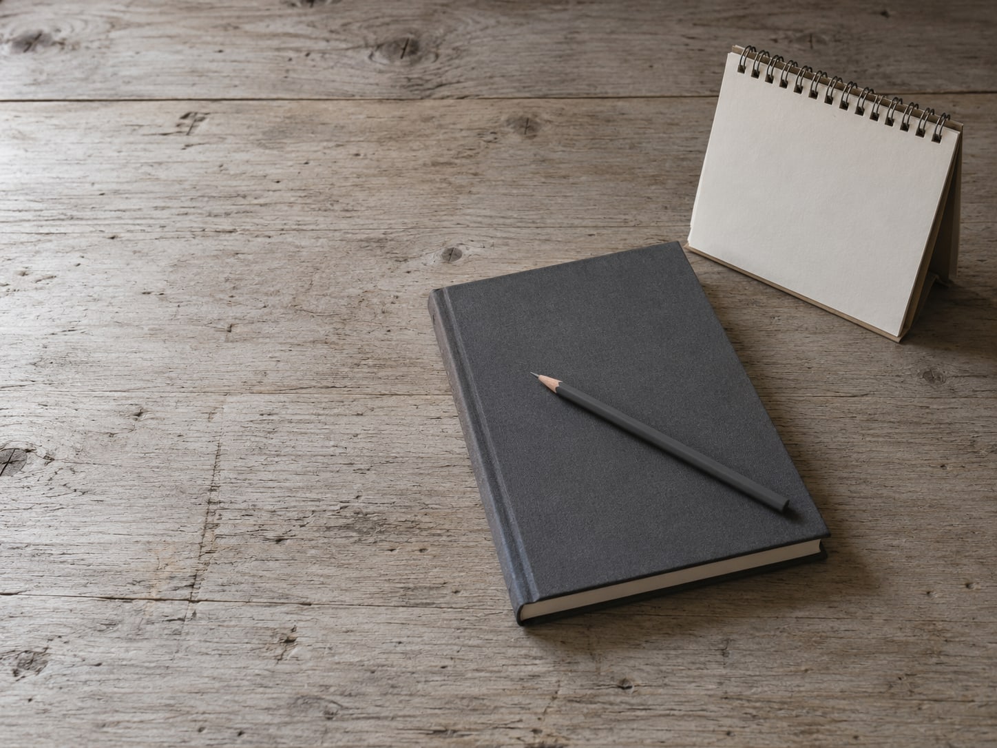 A closed notebook beside a small wall calendar on a wooden desk, with a pencil laid across the cover.