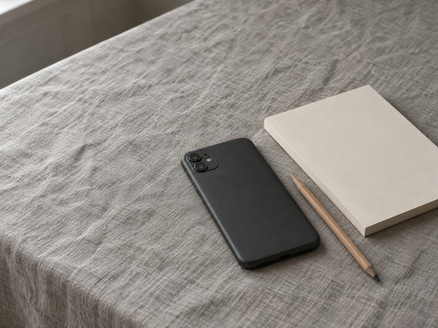 A phone lying face down next to a pencil and a blank notepad on a gray linen tablecloth.
