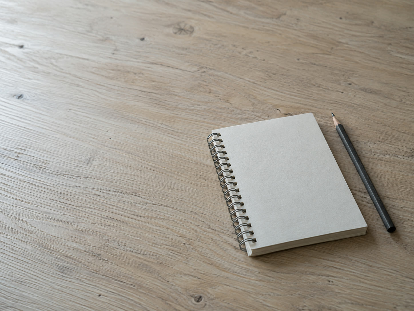 Un cuaderno espiral cerrado sobre una mesa de roble vacía junto a un lápiz afilado, iluminado por luz suave de ventana.