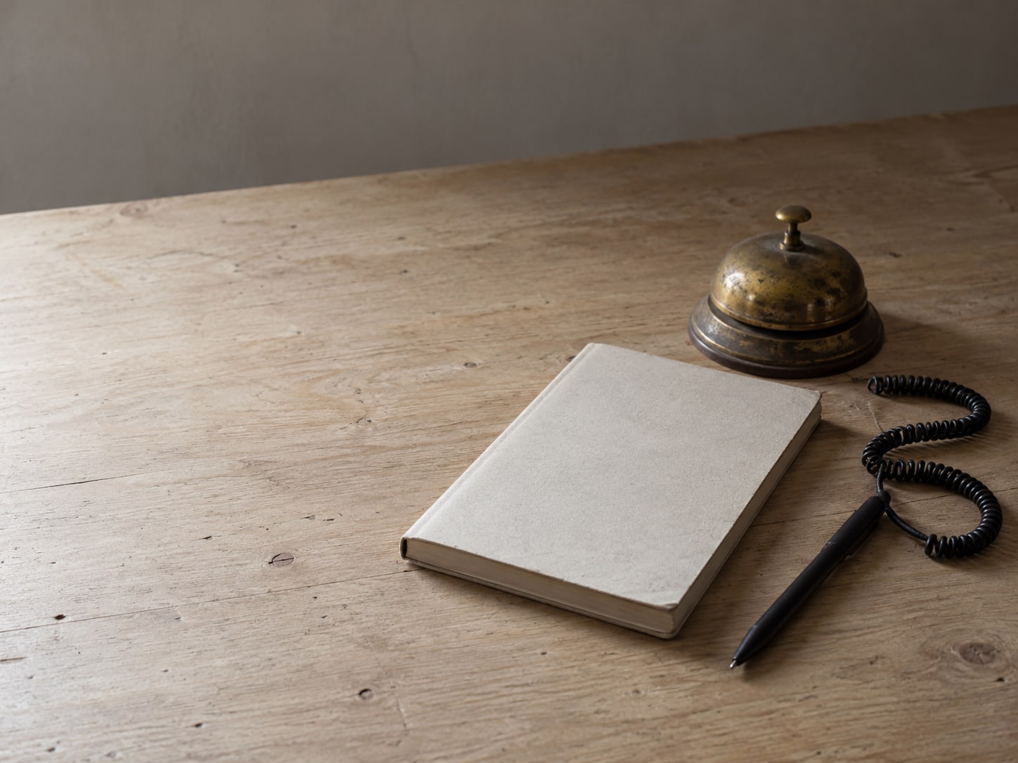 A closed notebook beside a brass desk bell and a coiled pen on a pale wooden counter.