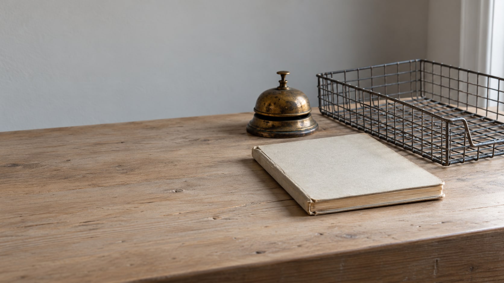 A closed notebook and a brass desk bell beside an empty wire inbox tray on a pale wooden counter.