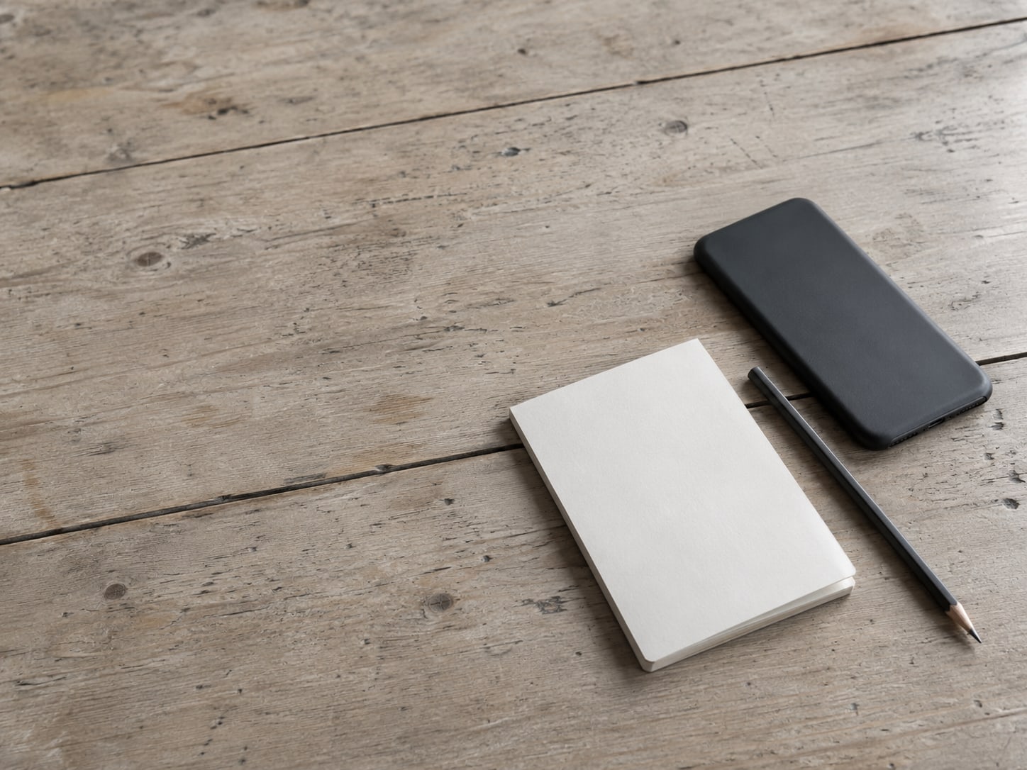 A closed phone lying face down beside a blank notepad and a pencil on a wooden counter.