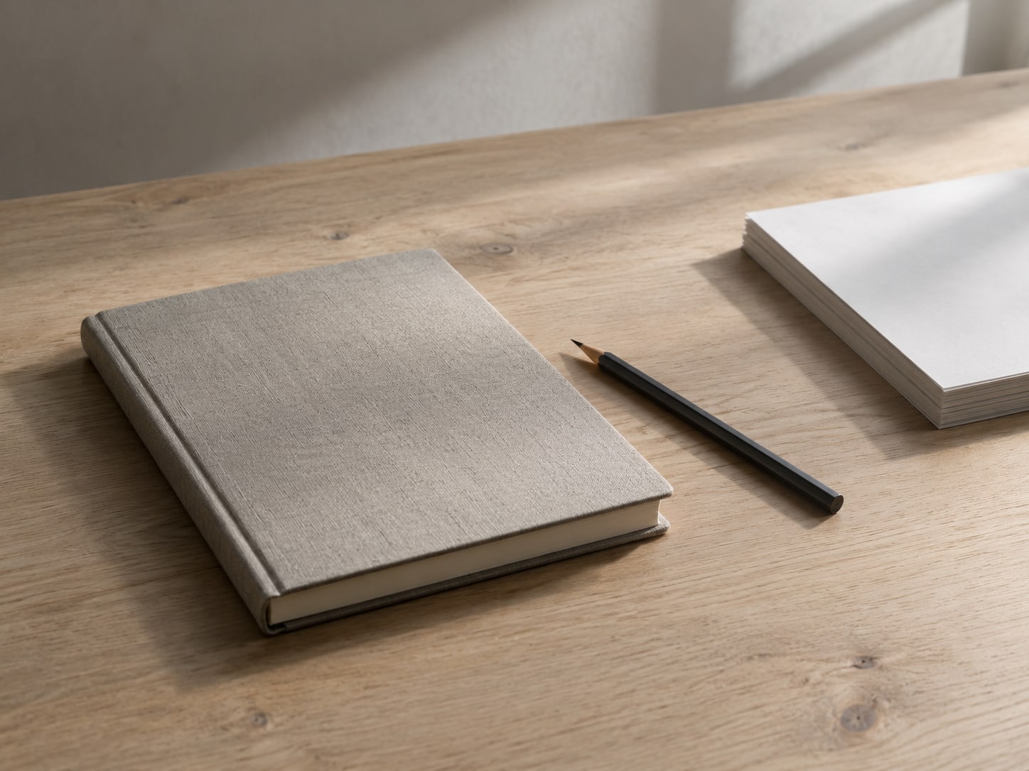 A closed notebook and a pencil resting on a clean wooden desk beside a small stack of blank paper.