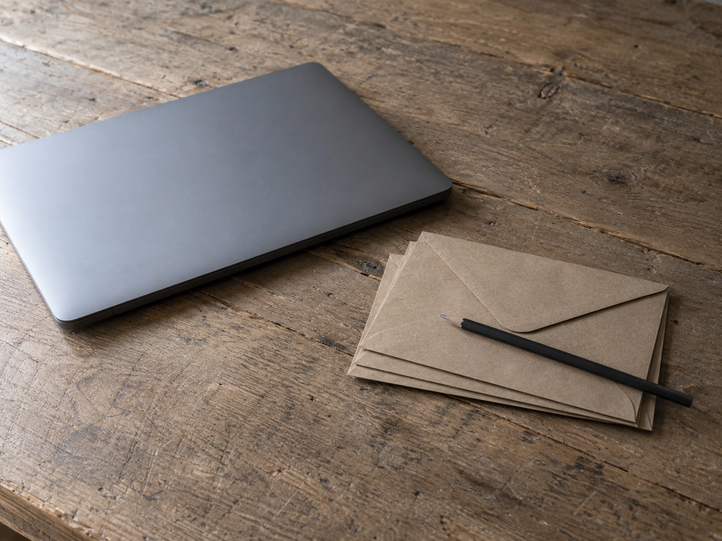 A closed laptop beside a small stack of sealed envelopes on a worn wooden desk, with a pencil lying across the top envelope.