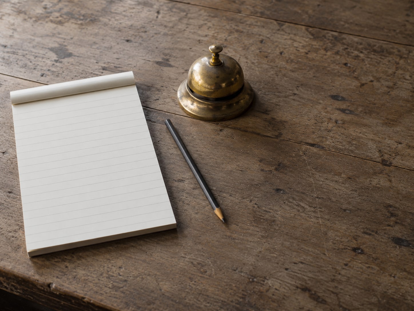 A pencil and a blank lined notepad on a wooden desk beside a small brass desk bell.