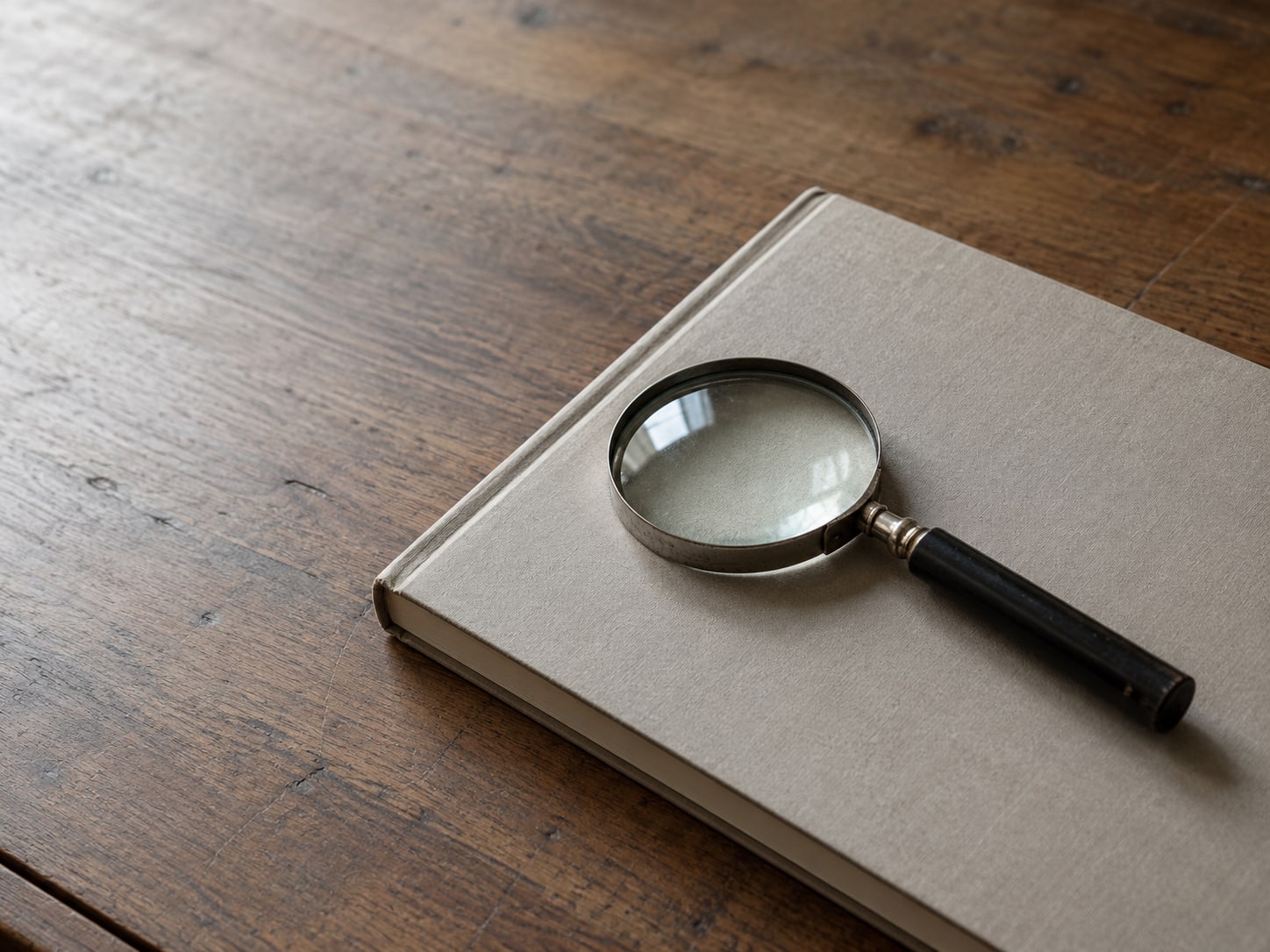 A magnifying glass lying on a closed paper ledger on a wooden desk.