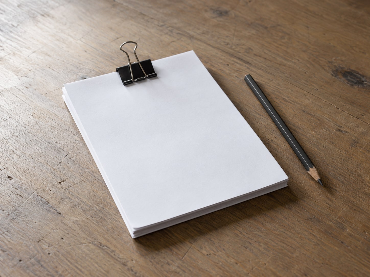 A short stack of blank typing paper held by a metal binder clip next to a pencil on a wooden desk.
