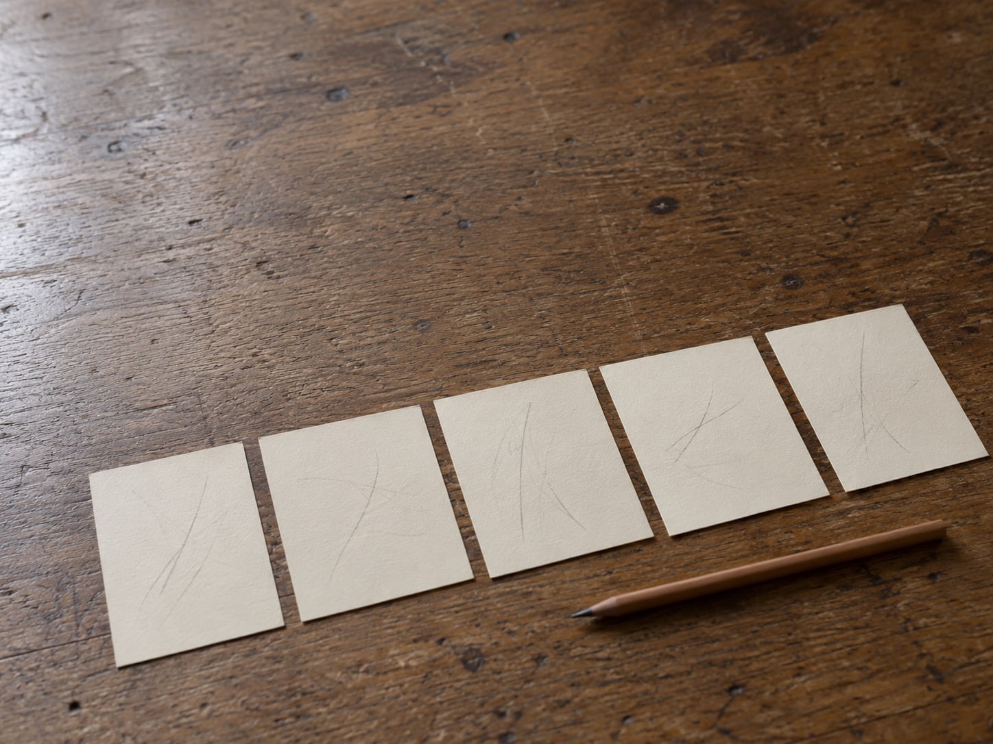 Five blank index cards in a row on a desk, each with a few faint pencil marks and a pencil lying beside them.