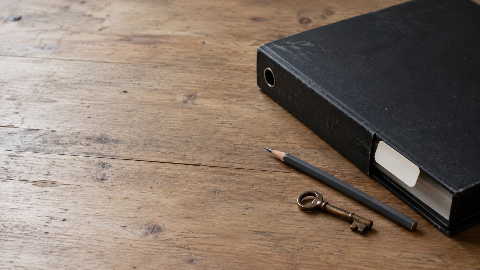 Una carpeta negra cerrada con una pestaña de índice en blanco sobre un escritorio de madera limpio, junto a un lápiz afilado y una pequeña llave de latón.
