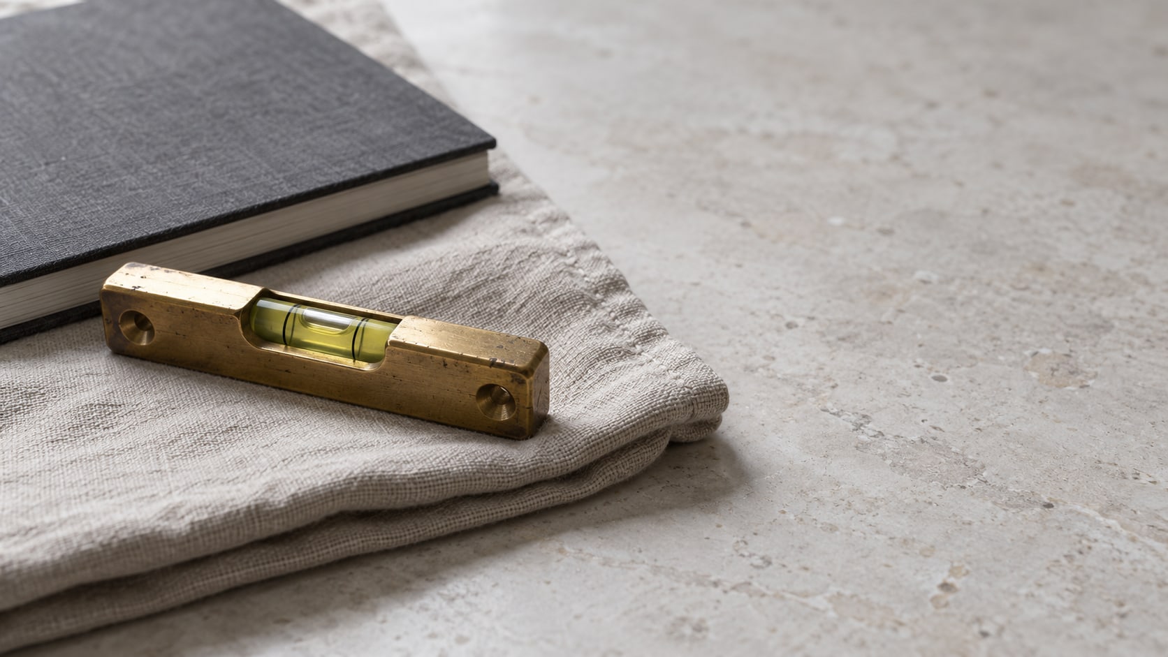 A small brass spirit level lying on a folded linen cloth beside a closed notebook on a pale stone surface.