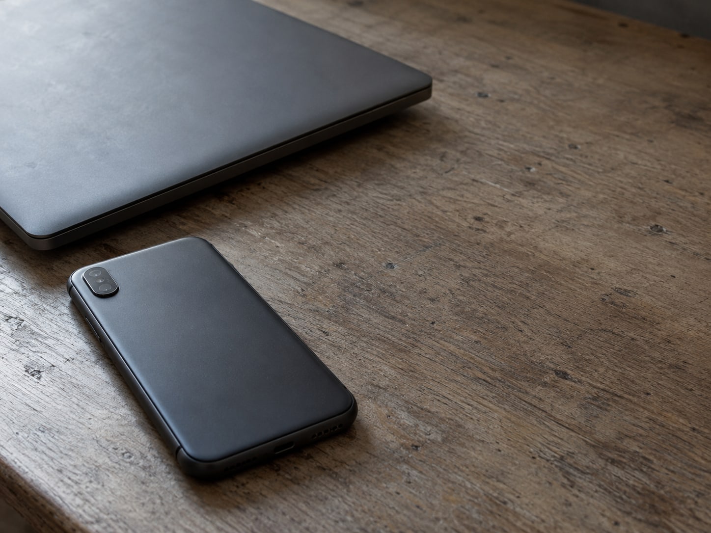 A smartphone lying face down beside a closed laptop on a plain wooden desk.