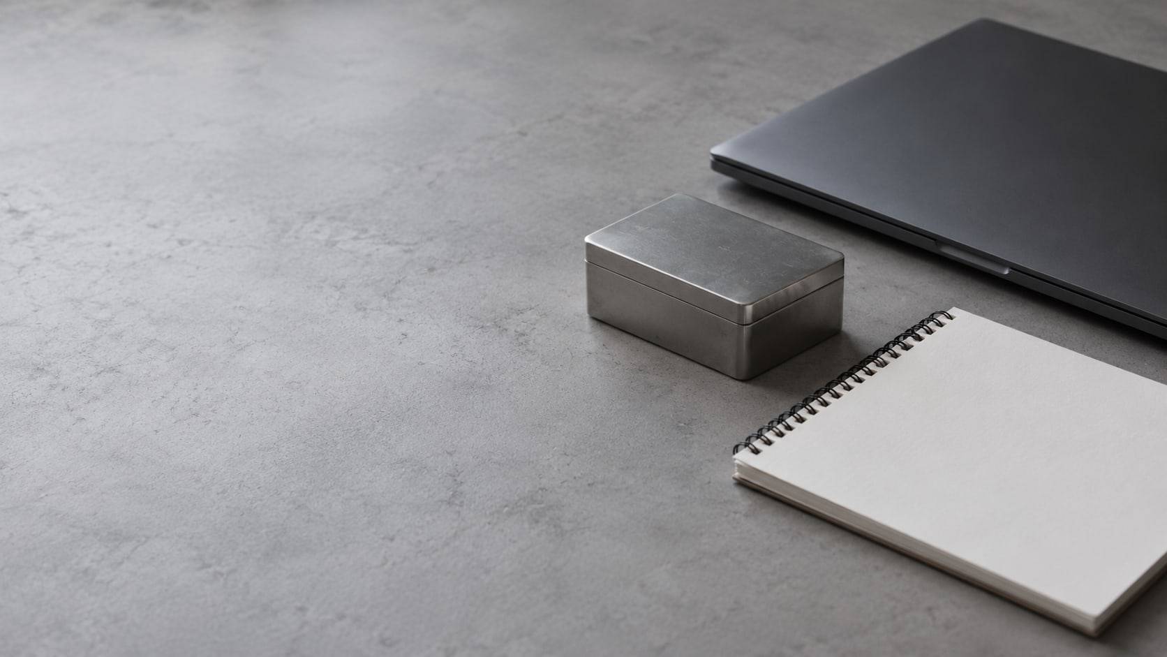 A small steel box beside a closed laptop and a blank notepad on a clean gray desk.