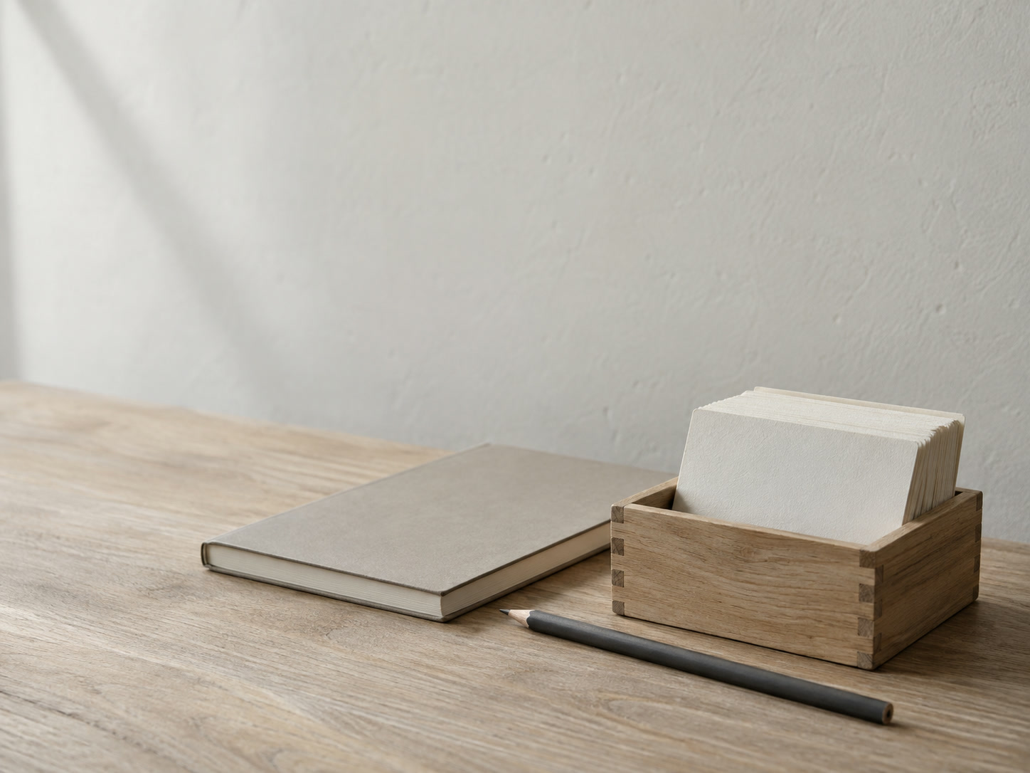 A small wooden box of blank index cards beside a closed notebook and a pencil on a pale oak desk.