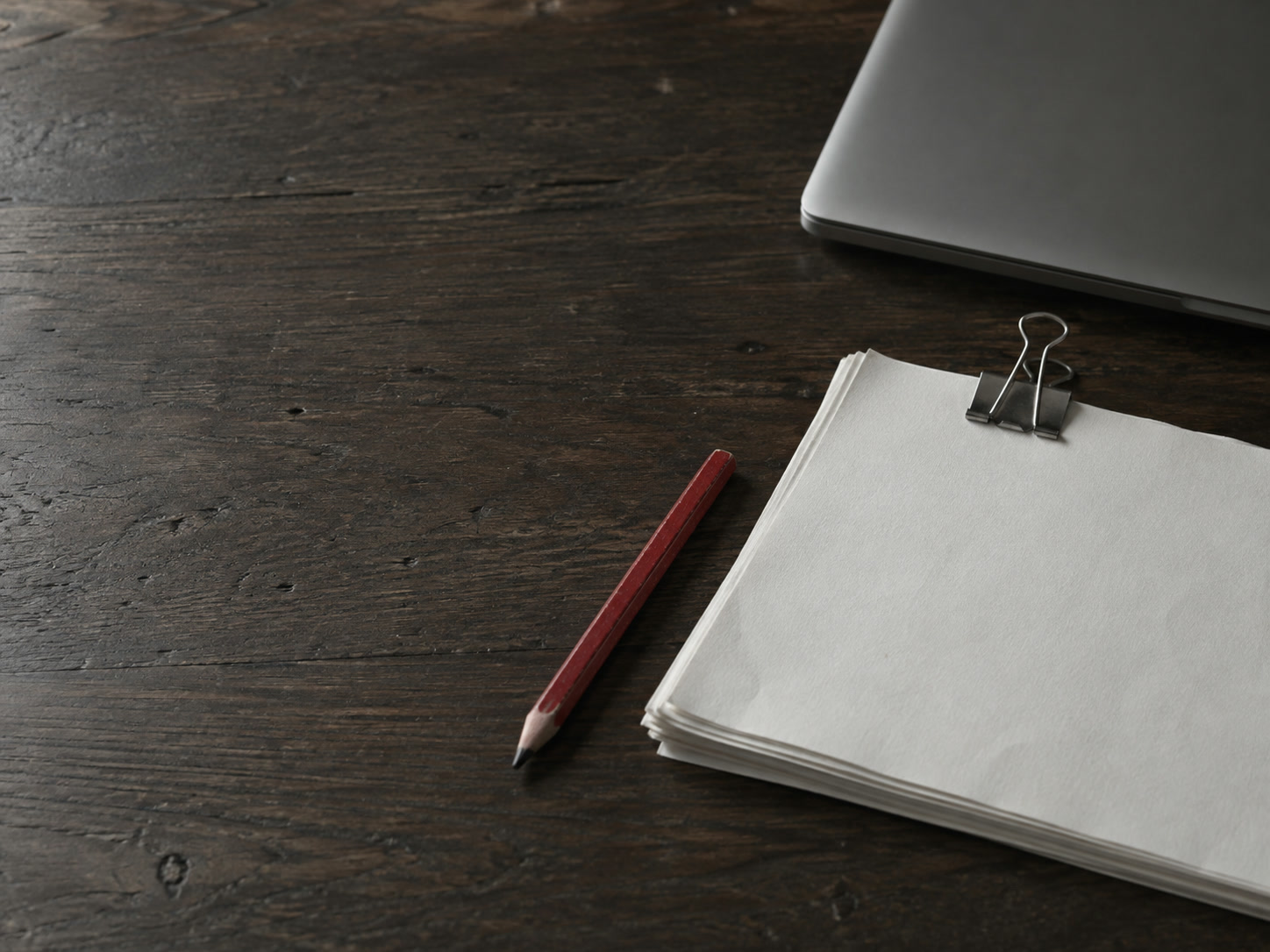 A thin stack of blank paper held with a binder clip beside a red pencil and a closed laptop on a walnut table.