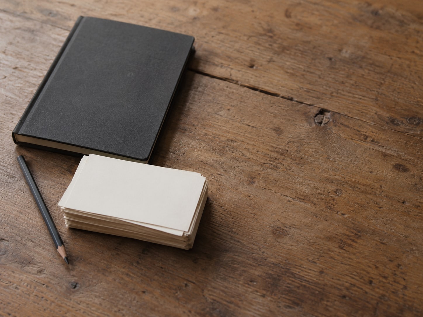 A closed notebook, a pencil and a stack of blank index cards laid out on a worn wooden desk.