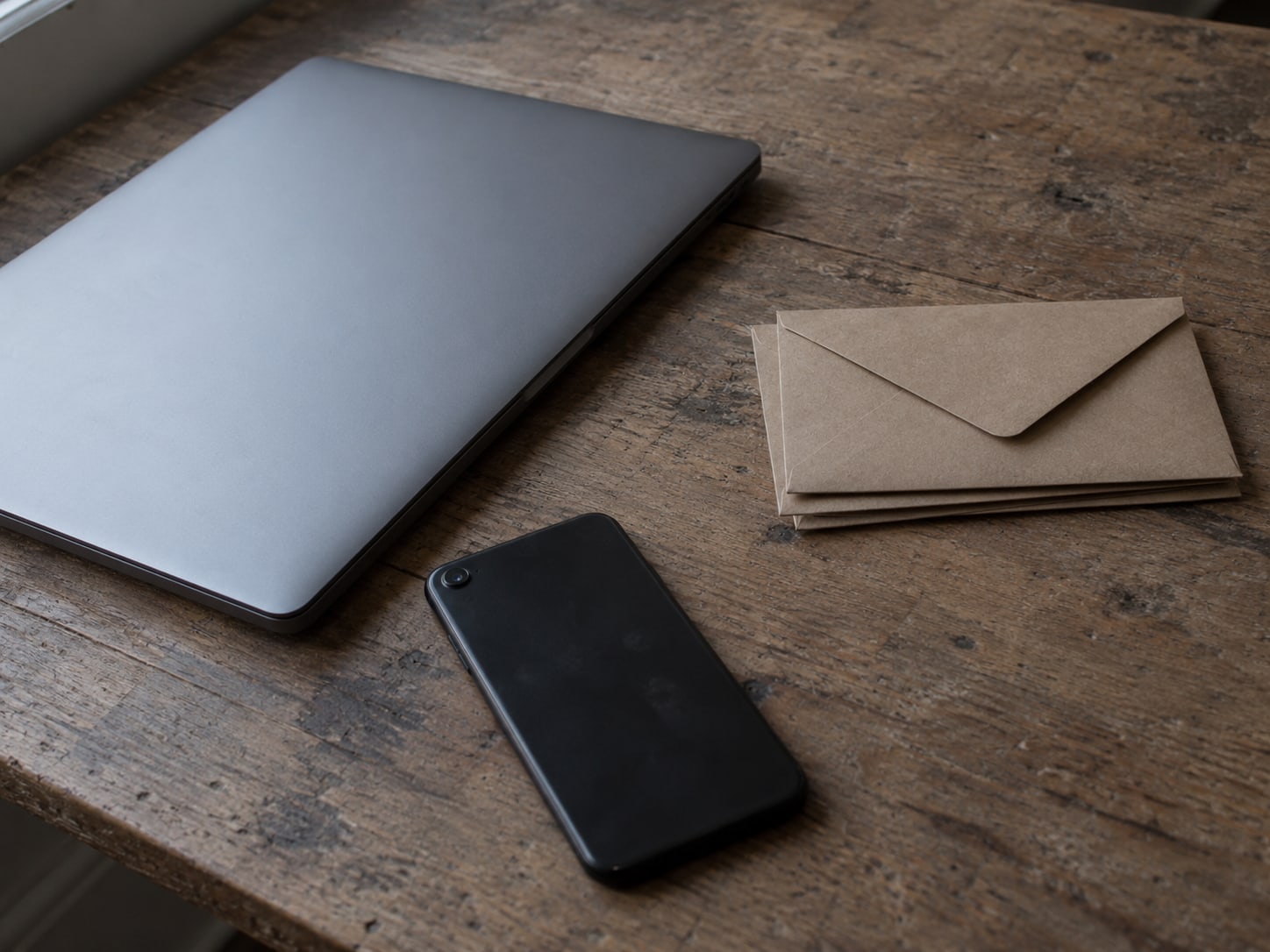 A closed laptop and a phone lying face down beside a small stack of unopened mail on a wooden desk.