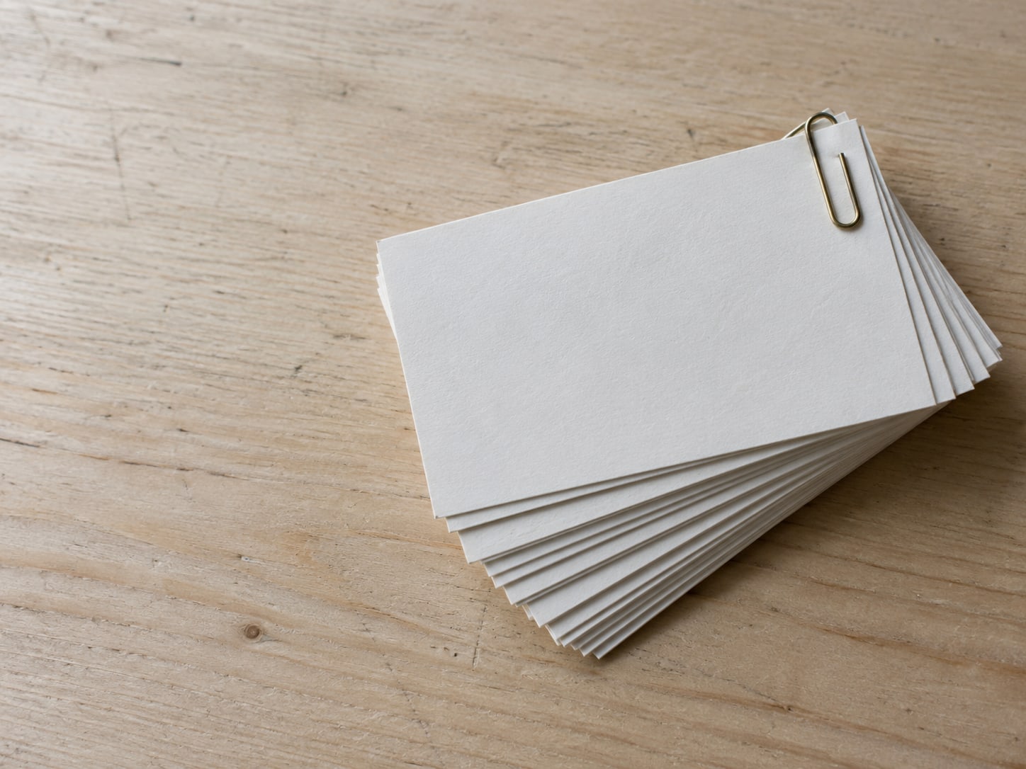 Un abanico de tarjetas de índice en blanco sujetas con un clip de latón sobre un escritorio de madera clara.