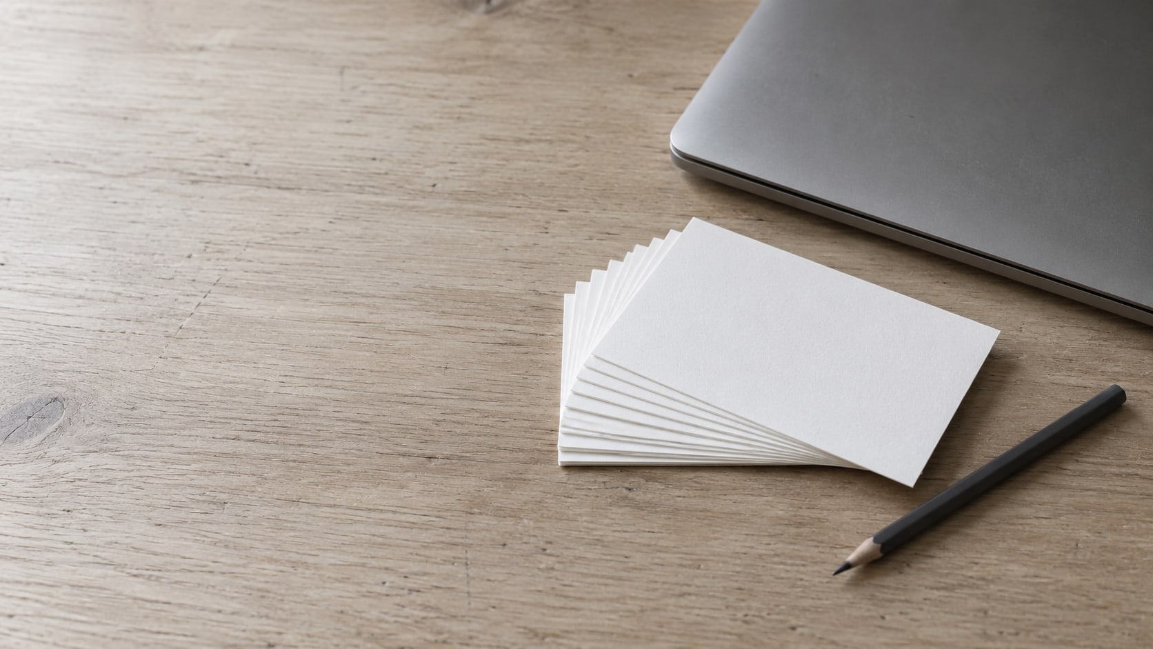 A fanned stack of blank index cards beside a closed laptop and a pencil on a pale wooden desk.