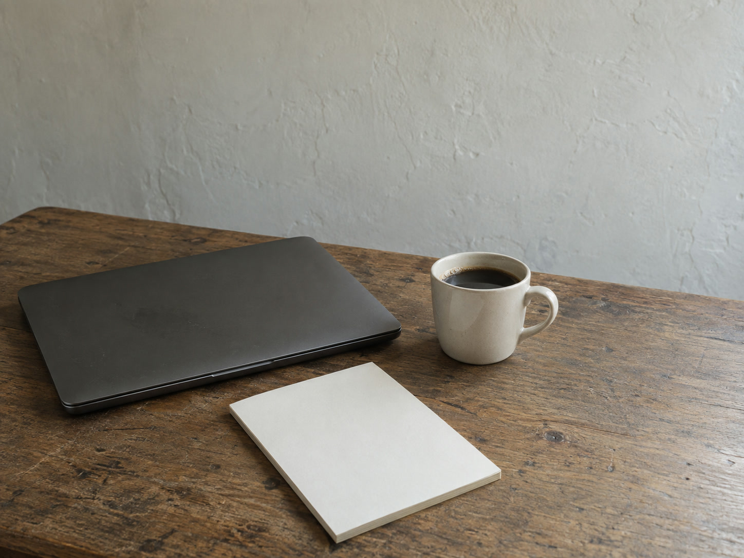 A closed laptop beside a mug of coffee and a blank notepad on a wooden desk in early morning light.