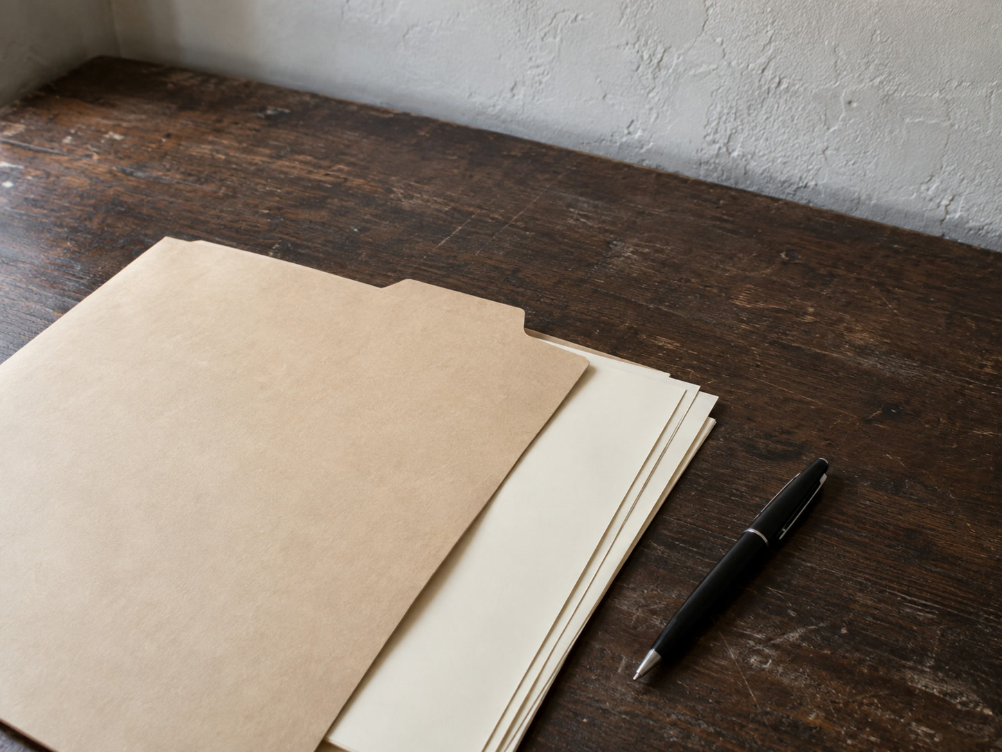 A plain manila folder holding a few blank sheets and a pen on a dark wooden desk.