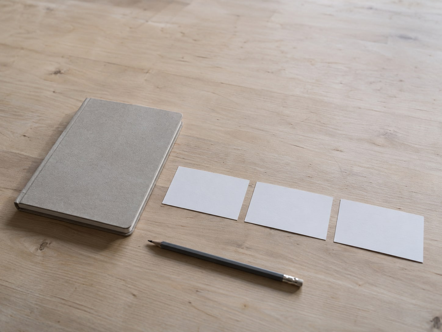A closed notebook beside three small blank index cards lined up on a pale wooden desk.