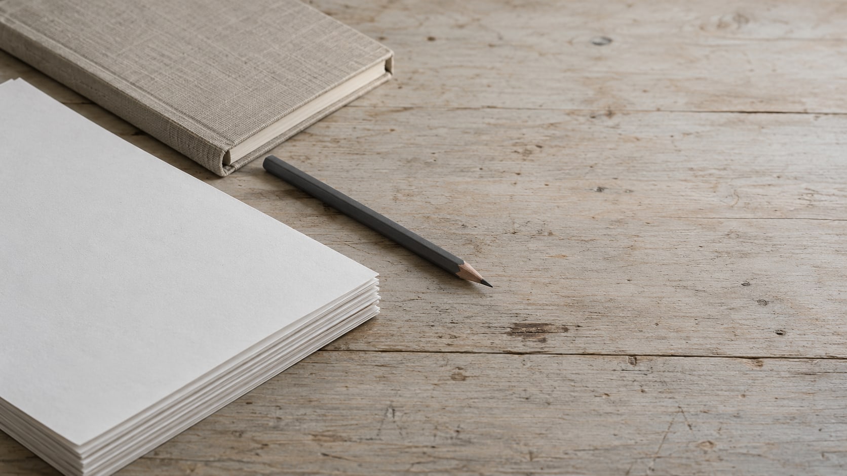 A short stack of printed pages with matching corners aligned on a wooden desk beside a closed notebook and a pencil.