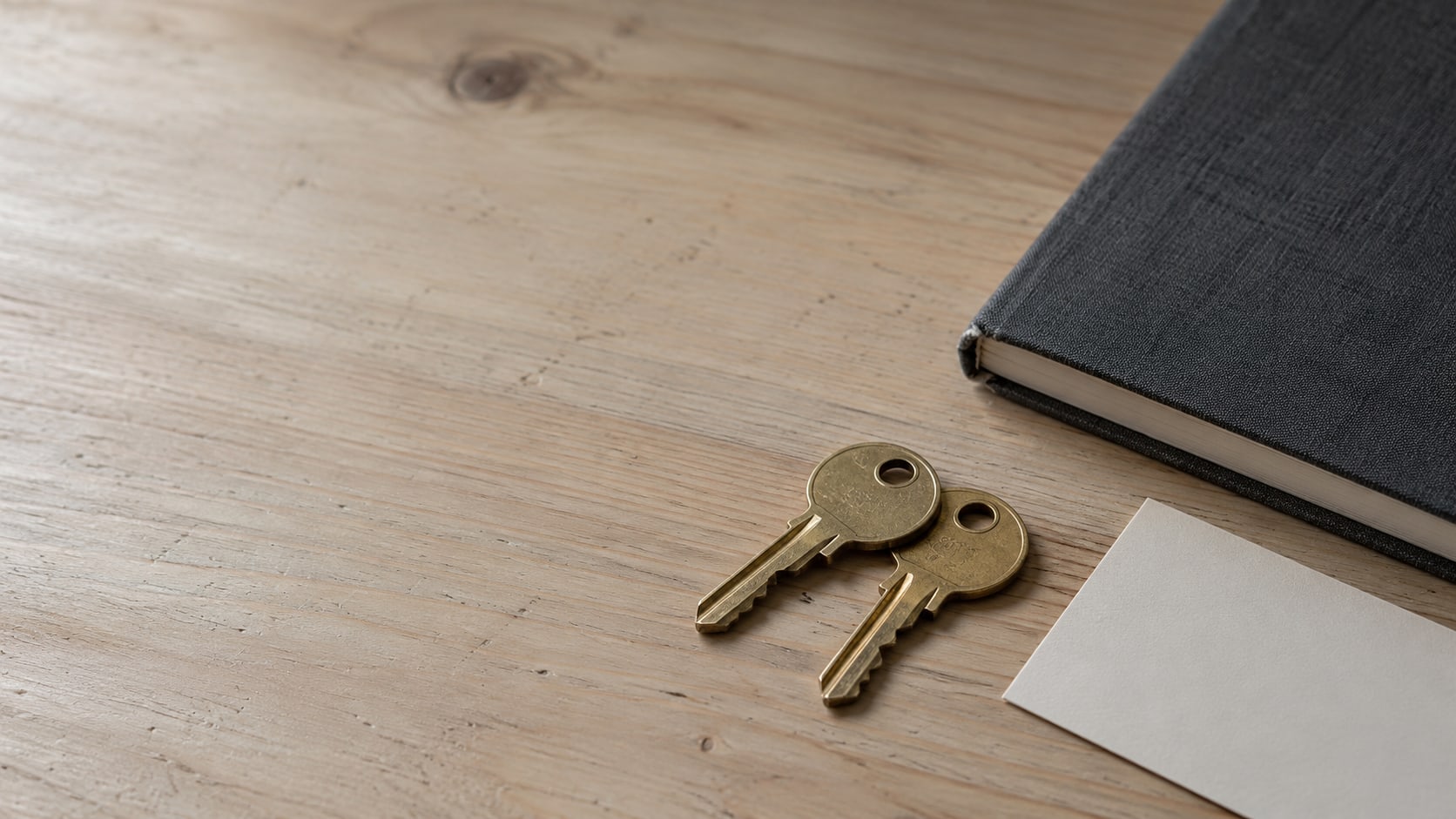 Two matching brass keys on a plain wooden desk beside a closed notebook and a blank index card.