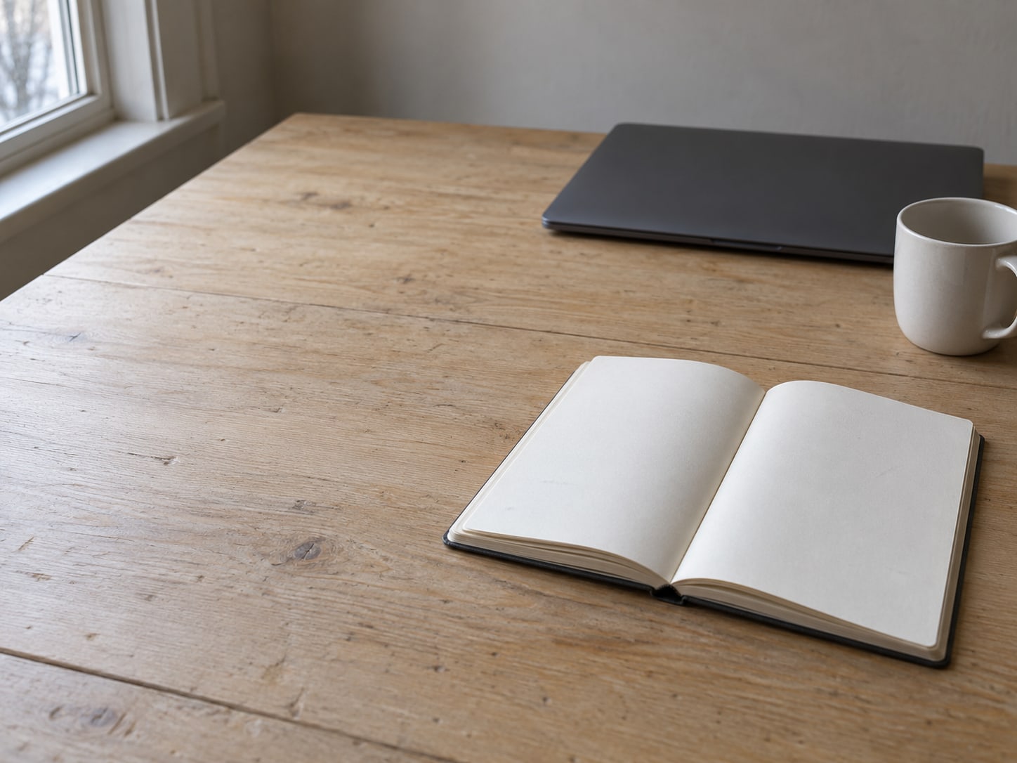 Una libreta en blanco abierta con marcas de lápiz tenues, junto a una laptop cerrada y una taza de cerámica sobre una mesa de madera.