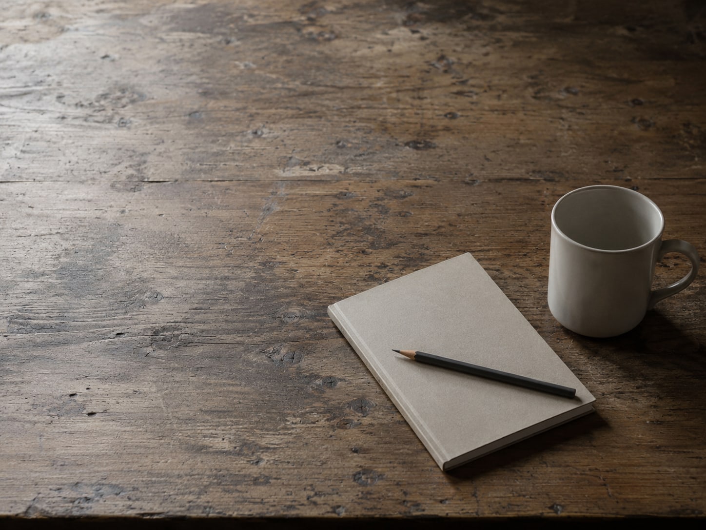 A wooden desk with a single sharpened pencil resting on a closed notebook beside an empty coffee mug.