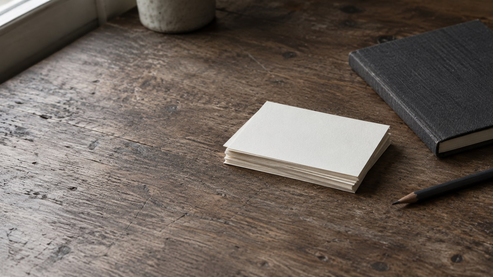 A small stack of blank cards beside a closed notebook and a pencil on a wooden desk.