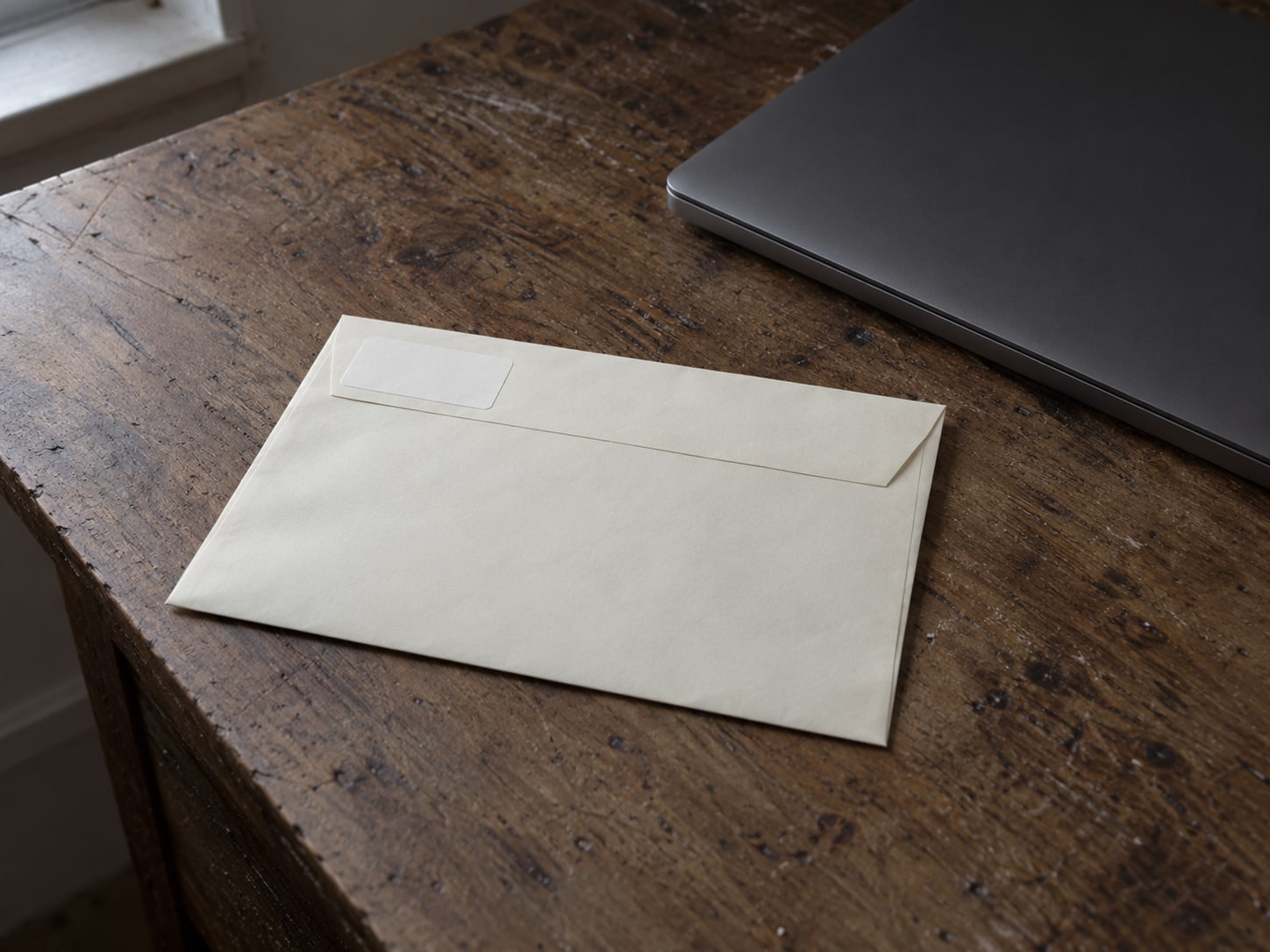 A neatly addressed envelope with a blank return label sits beside a closed laptop on a worn desk.