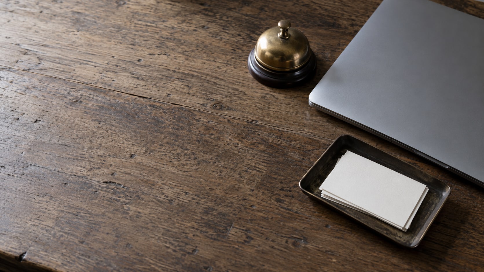 A wooden desk seen from above with a closed laptop, a brass desk bell and a small tray holding a few blank index cards.