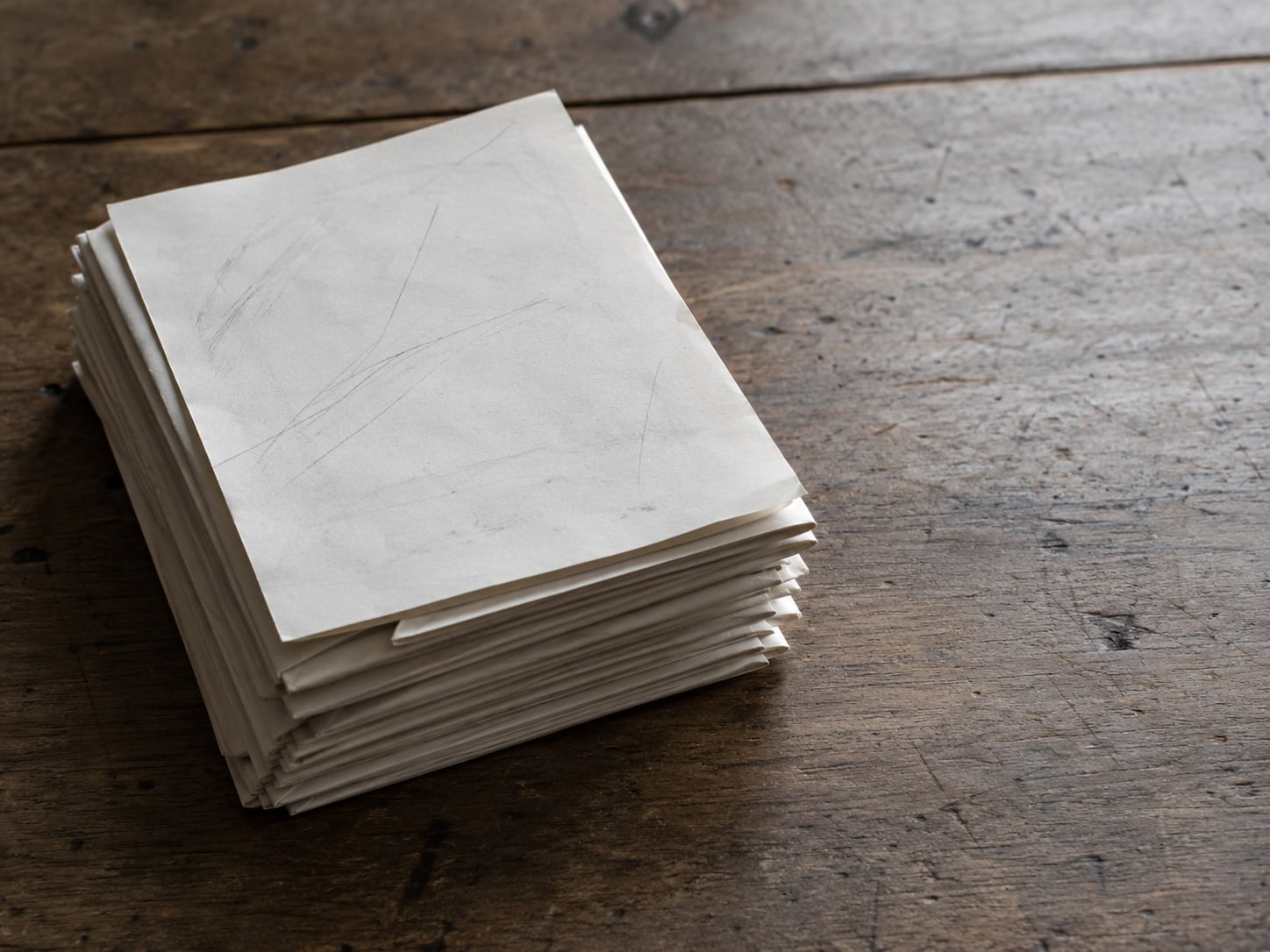 A single sheet of blank paper with faint pencil marks lying on top of a thick stack of folded letters on a wooden table.