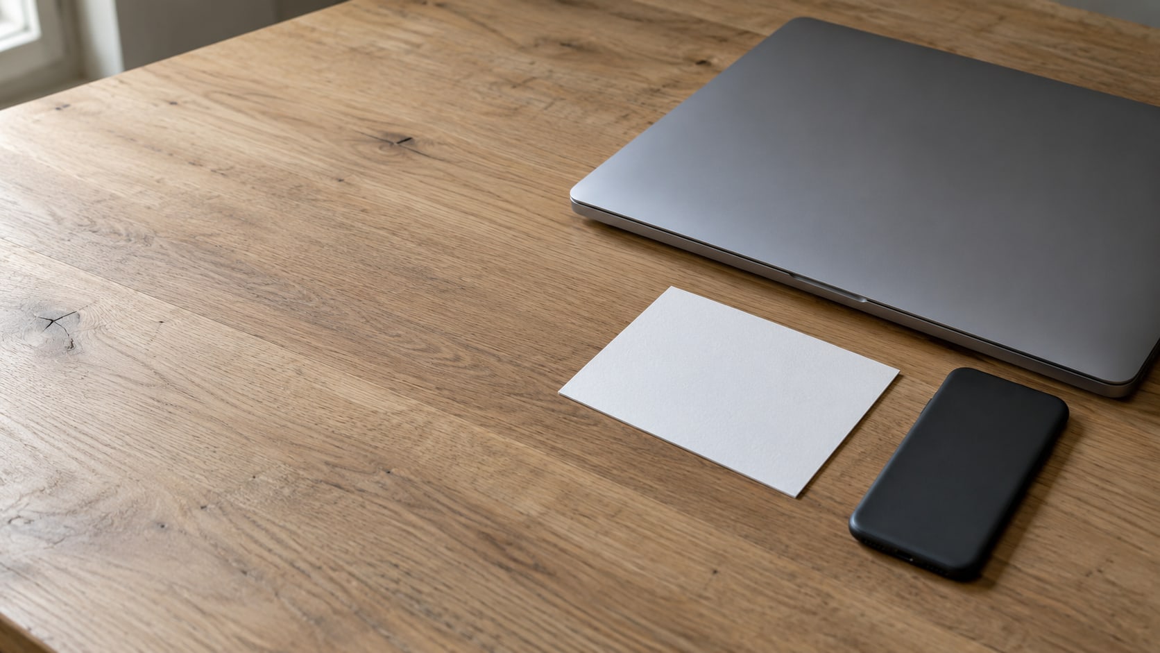 A blank index card and a closed laptop on a clean wooden desk, with a phone lying face down beside them.
