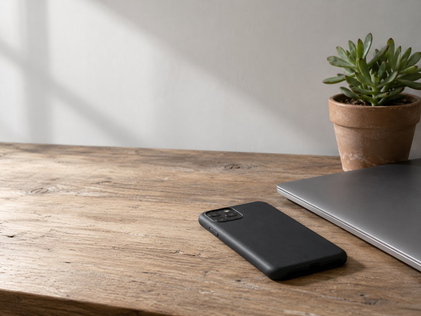 Un teléfono boca abajo sobre una mesa de madera, junto a una computadora portátil cerrada y una pequeña suculenta en maceta.