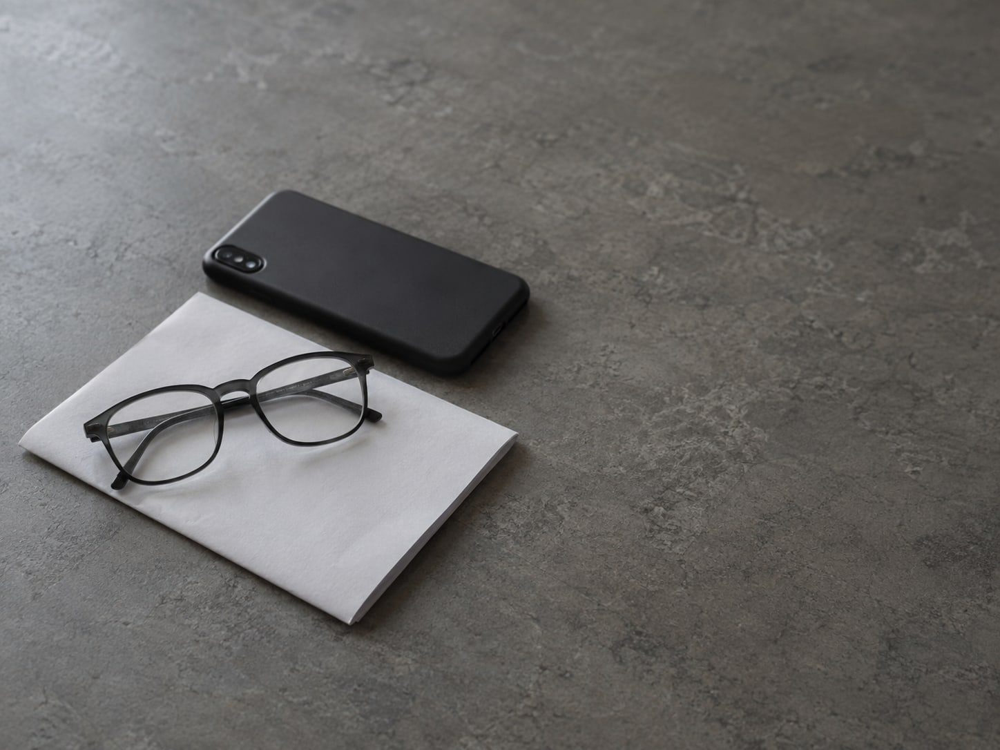 A pair of reading glasses resting on a folded sheet of blank paper next to a switched-off phone lying face down.