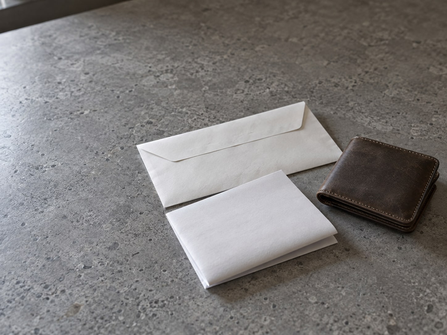 An unopened envelope and a folded bank statement with blank faces resting beside a closed wallet on a gray stone countertop.