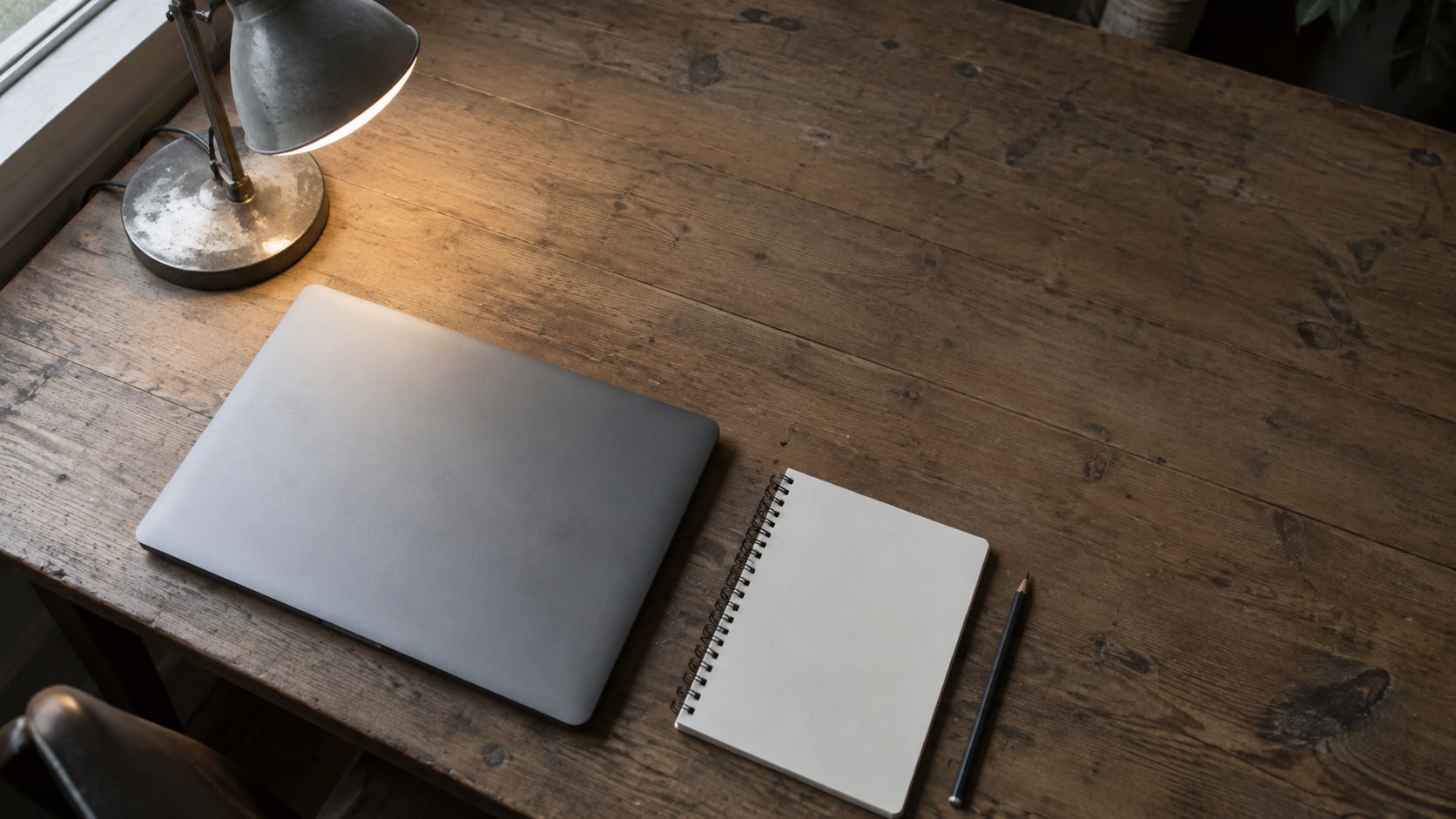 A closed laptop, a blank notepad and a pencil on a plain wooden desk beside a lit desk lamp, photographed from above.