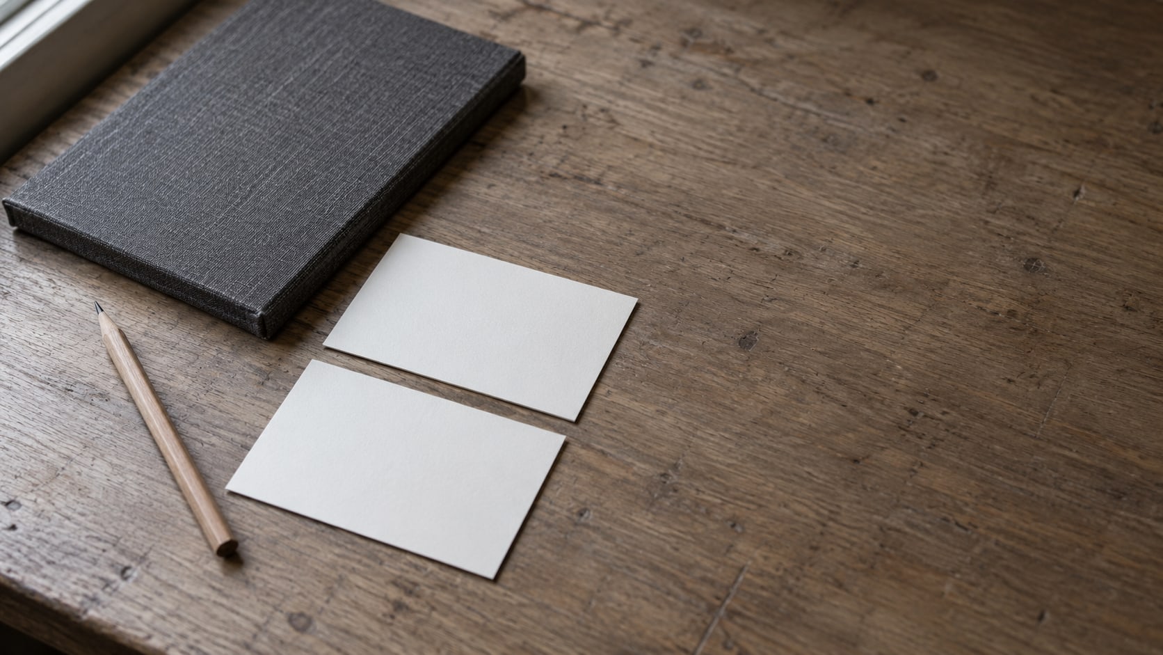 Two matching blank index cards and a pencil resting on a plain wooden desk beside a closed notebook.