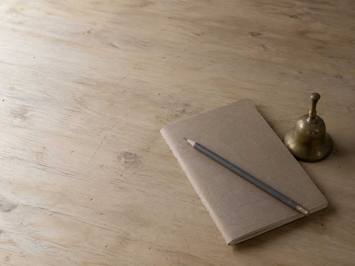 A closed notebook with a pencil resting across it on a clean wooden desk beside a small brass bell.