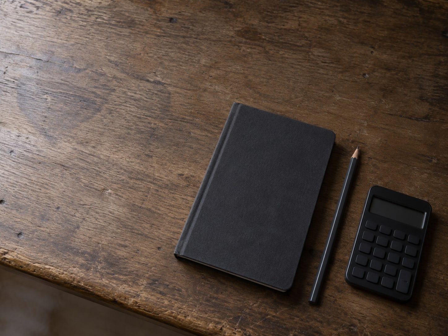A closed notebook and a pencil beside a small switched-off calculator on a worn wooden desk.