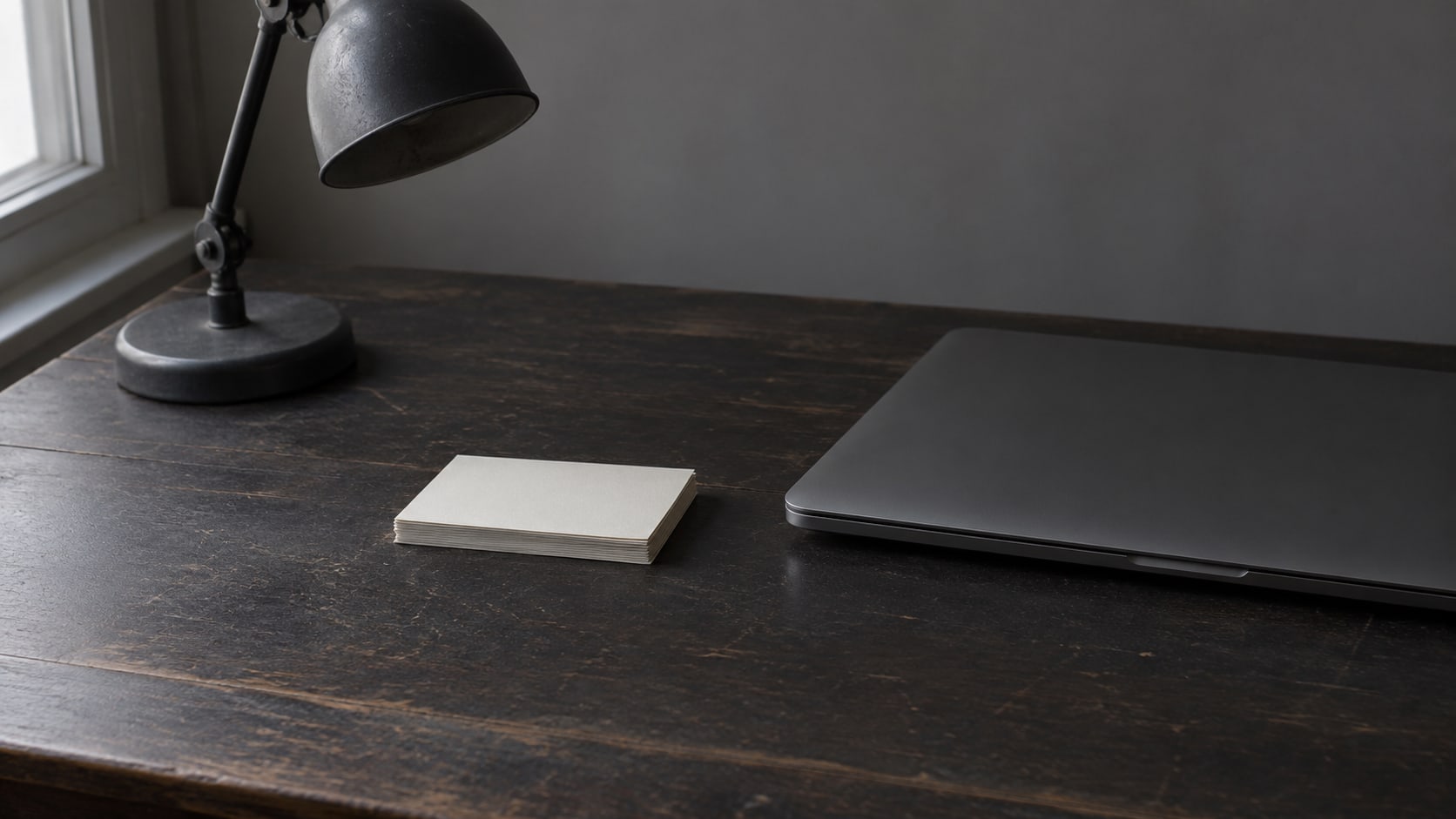 A closed laptop and a small stack of blank index cards beside a switched-off desk lamp on a dark wooden desk.