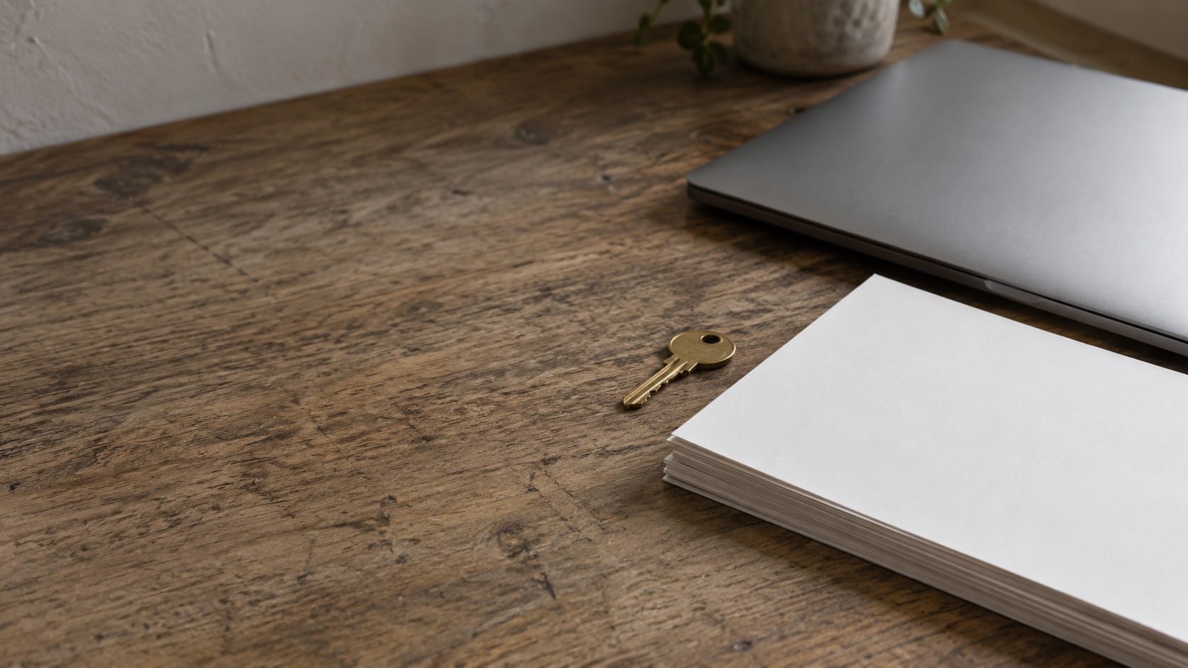 Una laptop cerrada y una pila de cotizaciones en papel en blanco junto a una pequeña llave de latón sobre un escritorio de madera gastado.