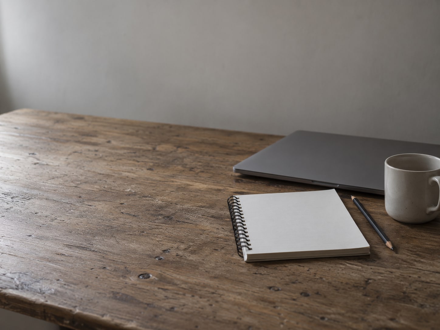 Un bloc de espiral en blanco con un lápiz junto a una laptop cerrada y una taza sobre un escritorio.