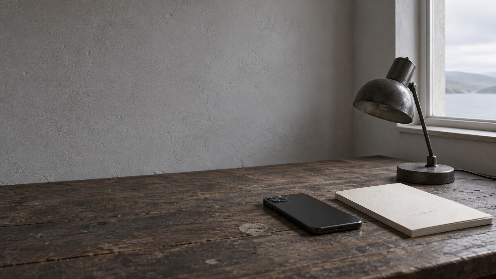 A face-down phone beside an unlit desk lamp and a blank notepad with faint pencil marks on a dark desk.