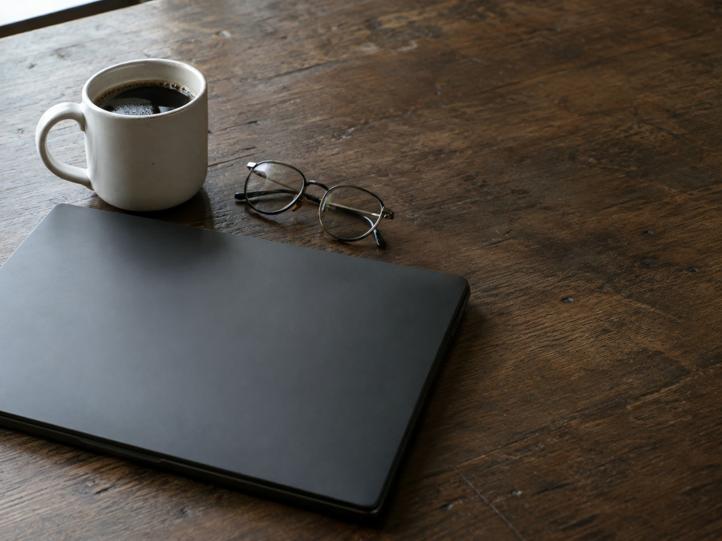 A closed laptop on a wooden desk beside a folded pair of reading glasses and a plain white coffee mug.