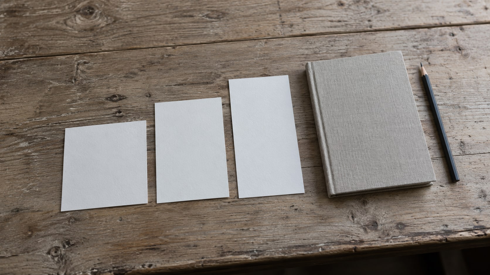 A row of blank square and vertical printed cards laid out on a light wooden desk beside a closed notebook and a pencil.
