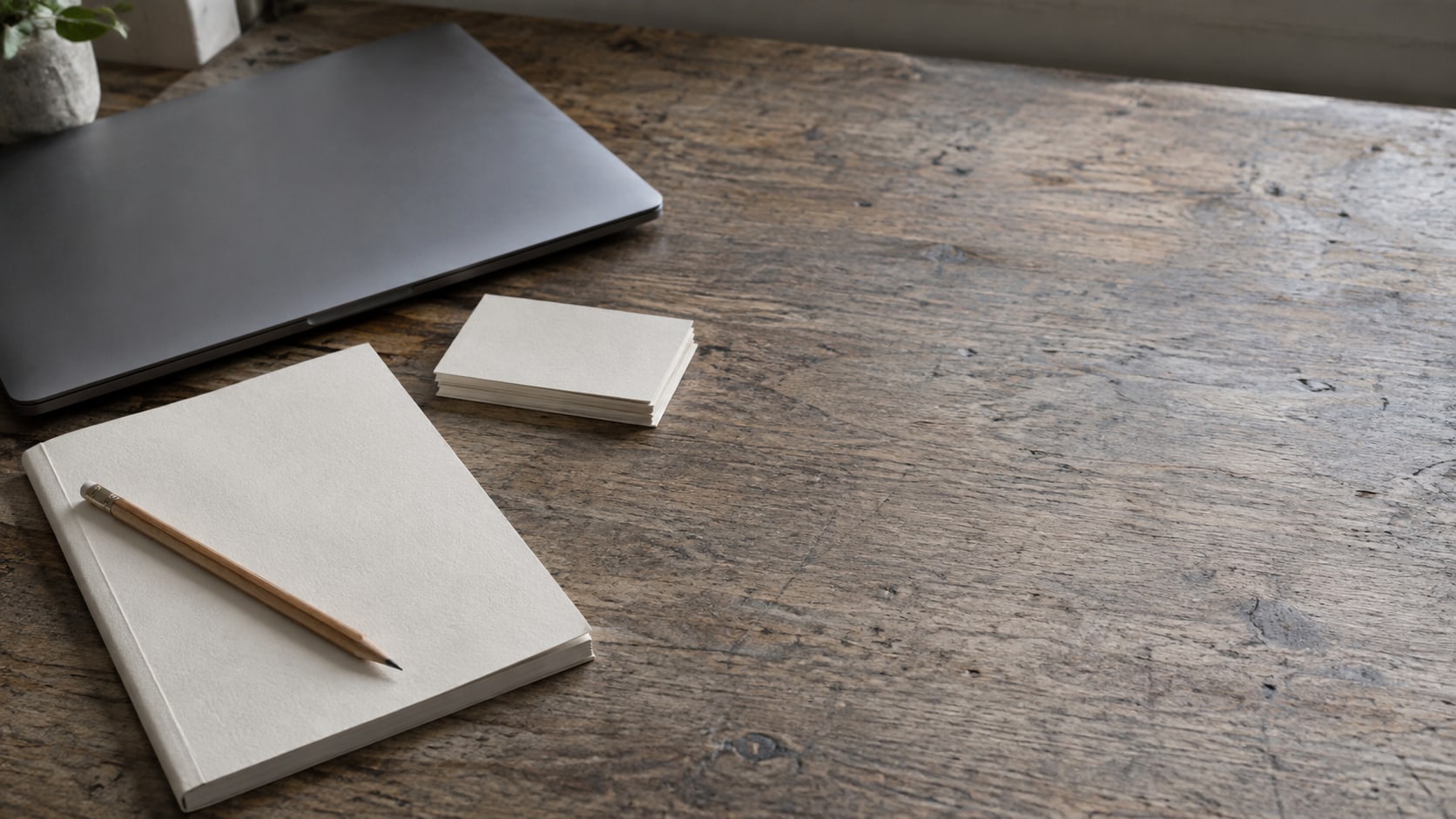A closed laptop, a blank notebook and a pencil arranged on a worn wooden desk beside a small stack of blank index cards.