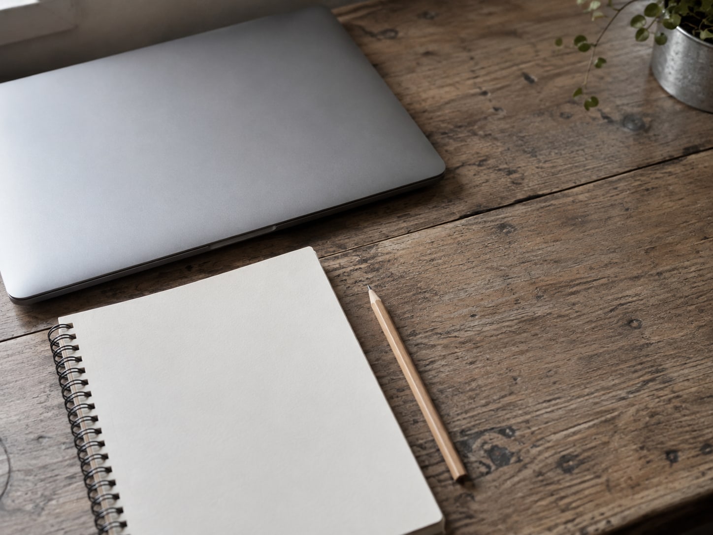 Una laptop cerrada junto a un cuaderno de papel en blanco y un lápiz afilado sobre un escritorio de madera gastada.