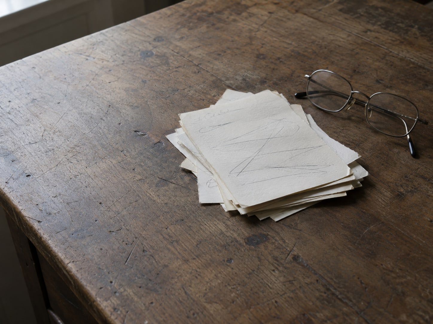 A small pile of paper slips with unreadable pencil scribbles and a few crossed out lines, next to a pair of reading glasses.
